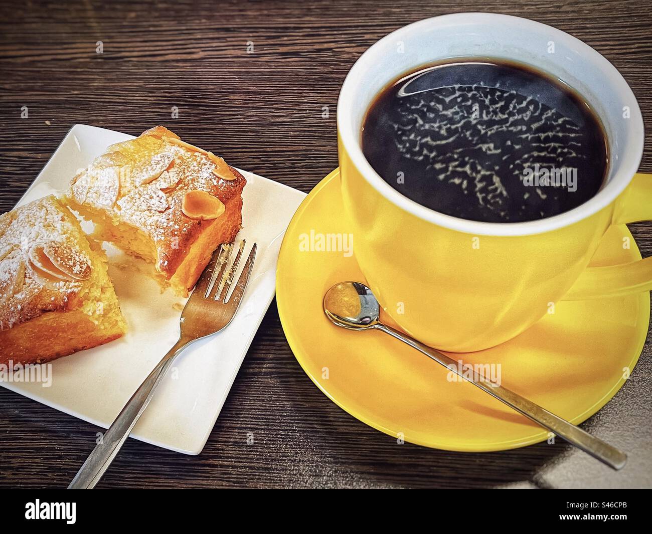 Schwarzer Kaffee/langer schwarzer Kaffee in einer gelben Kaffeetasse mit Untertasse und einer Scheibe glutenfreiem orangen Kuchen auf weißem Teller auf Holztisch. Snack, Kaffeepause, Teestunde. Stockfoto