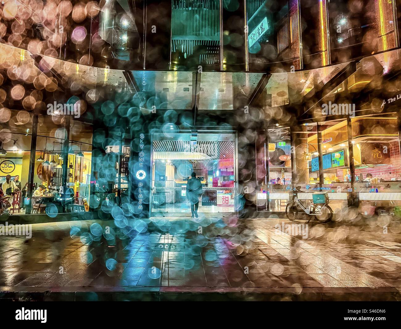 Blick durch nasses Glas auf einen Mann, der an einer nassen, regnerischen Nacht ein Restaurant in einem beleuchteten mehrstöckigen Gebäude betritt. Stockfoto