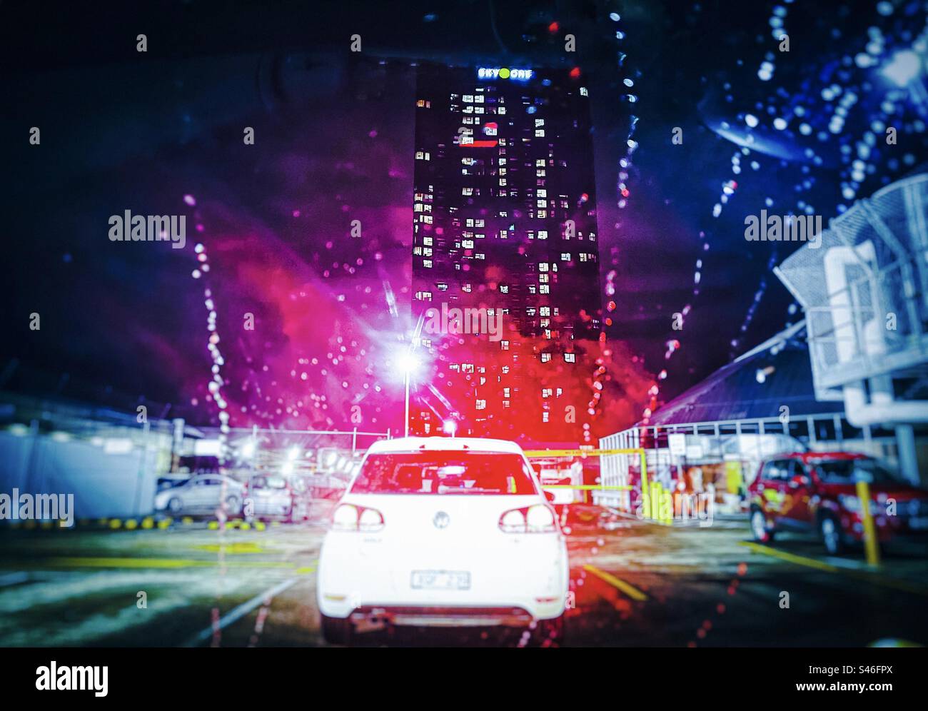 Beleuchteter Wolkenkratzer, Straßenlaterne, Gebäude, Autos und Neonlicht, das in einer regnerischen Nacht durch die nasse Windschutzscheibe gesehen wird. Farbiges Licht, Regentropfen, Unschärfe, Bewegung. - Smartphone-aufgenommenes Stockfoto