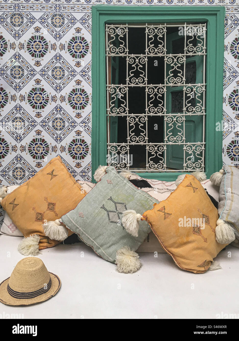 Stillleben in einem marokkanischen Riad mit bunten Wandfliesen, kunstvoll verzierten eisernen Fensterbezügen, Wurfkissen und Strohhut. Stockfoto