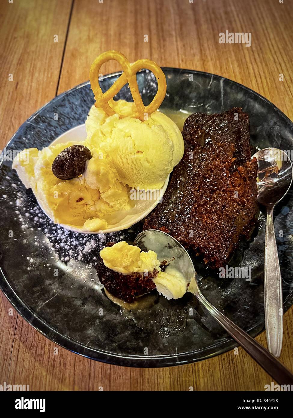 Blick aus einem hohen Winkel auf klebrigen Dattelpudding mit zwei Messlöffeln Eis, Brezel und Schokolade auf schwarzem Teller mit Löffeln auf dem Tisch. Stockfoto