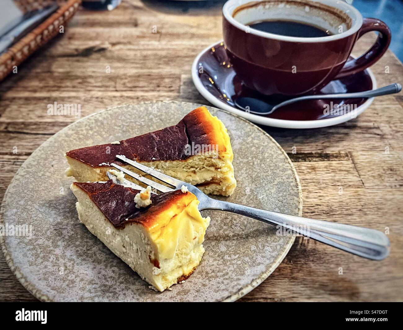 Blick aus einem hohen Winkel auf ein Stück baskischen Käsekuchen auf einem Teller mit Gabel und heißem Kaffee in einer Kaffeetasse mit Untertasse auf einem Holztisch im Café. Stillleben. Hintergründe. Verwöhnung. Versuchung. Stockfoto
