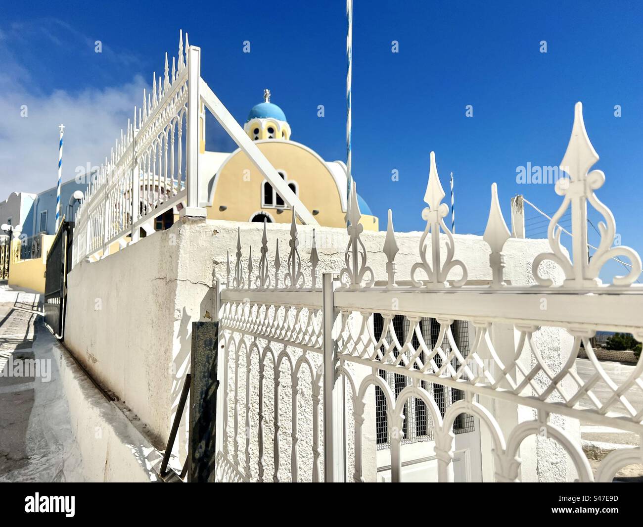 Reich verzierte weiß bemalte Tore und die gelbe, blaue Kuppelkirche des Heiligen Johannes, die russische enge Straße im Dorf Kontochori von Santorin. - Smartphone-aufgenommenes Stockfoto