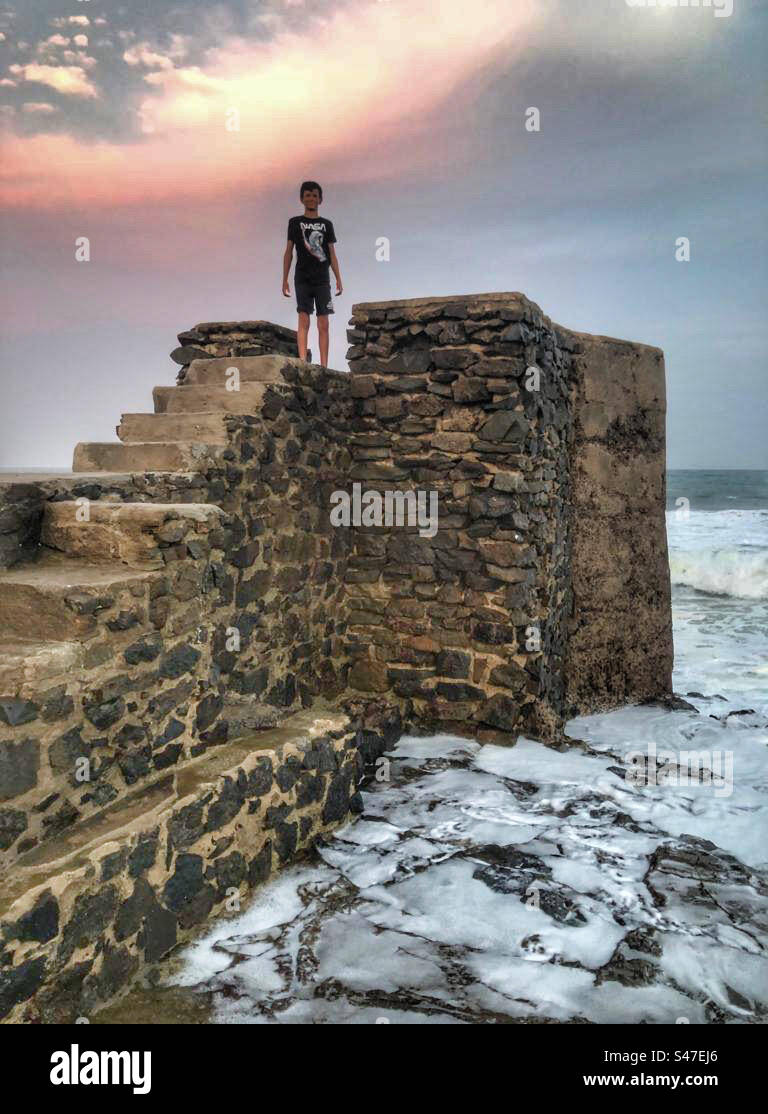 Ein kleiner Junge, der auf einer Steinstruktur steht, während der einfallenden Flut während des Sonnenuntergangs Stockfoto