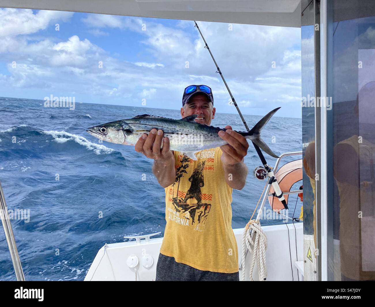 Fischer auf einem Boot, der stolz einen spanischen Makrelenfisch, eine Angelrute und ein Meer im Hintergrund hält Stockfoto