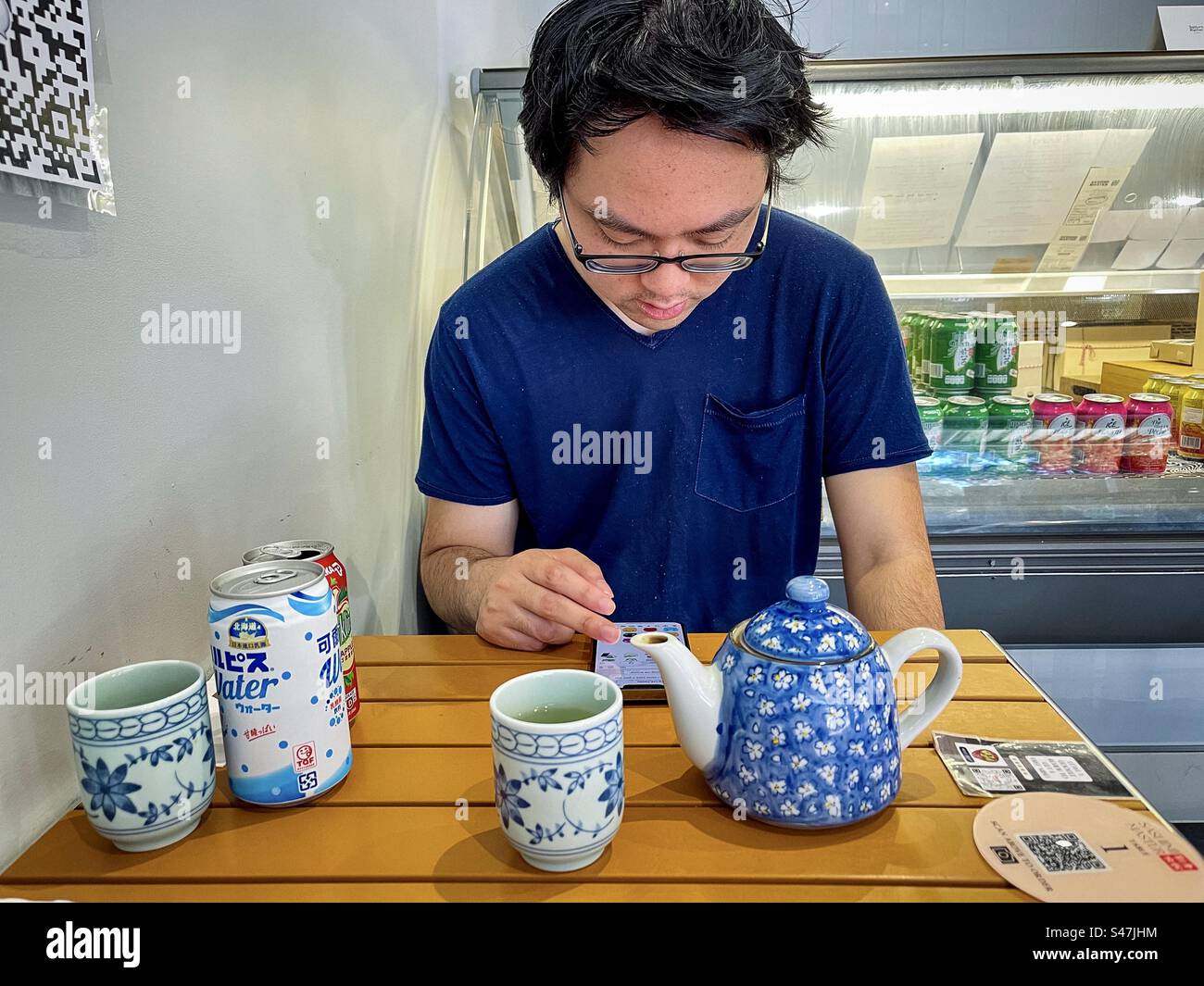 Junger asiatischer Mann in Brille mit QR/Quick Response Code und Handy, um Essen am Tisch in einem japanischen Restaurant zu bestellen. Wireless-Technologie. - Smartphone-aufgenommenes Stockfoto