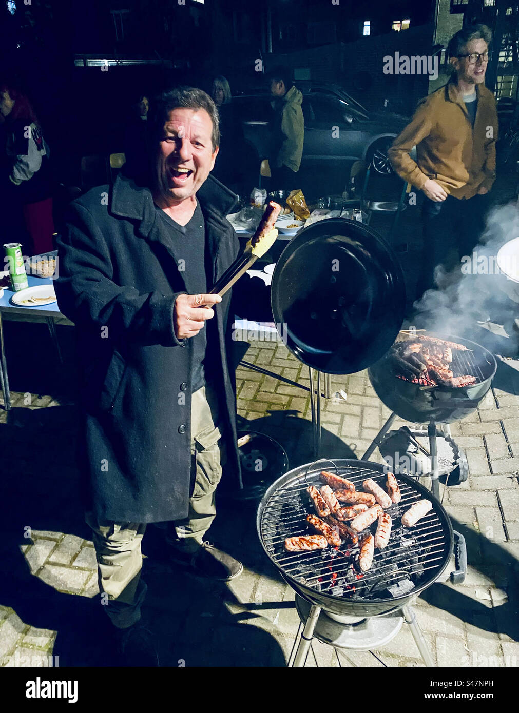 Ein Mann hält eine Wurst in einer Zange bei einem Barbecue am Abend in London - Smartphone-aufgenommenes Stockfoto