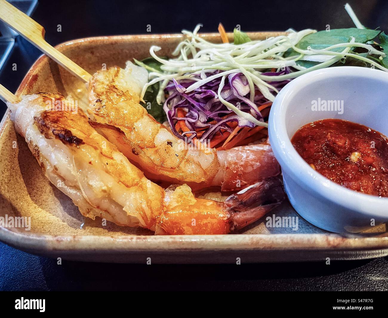 Nahaufnahme mit gegrillten Garnelenspießen und frischem Salat auf Teller mit einer Schüssel scharfer Chili-Sauce auf dem Tisch. Gesunde Ernährung. Asiatisches Essen. Würze. Sambal. - Smartphone-aufgenommenes Stockfoto