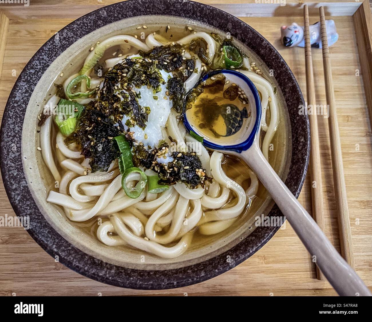 Direkt darüber, Nahaufnahme von Udon-Nudeln in Miso-Suppe mit Onsen Tamago/pochiertem Ei, Algen und Sesamsamen in Schüssel mit Löffel und Stäbchen auf Holztablett Washoku. Traditionelles japanisches Essen. - Smartphone-aufgenommenes Stockfoto