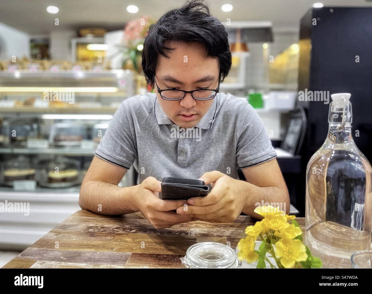 Junger asiatischer Mann mit Brille, der ein Handy benutzt, während er an einem Tisch in einem Café sitzt. Wireless-Technologie. Essen und Trinken. - Smartphone-aufgenommenes Stockfoto