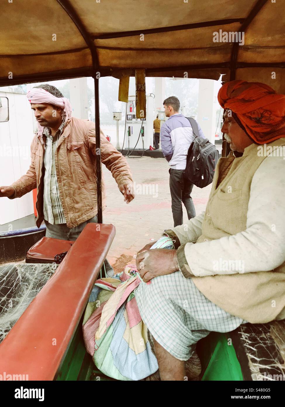 Ein älterer Mann mit rotem Turban sitzt in einer Autorikscha an einer Tankstelle - Smartphone-aufgenommenes Stockfoto