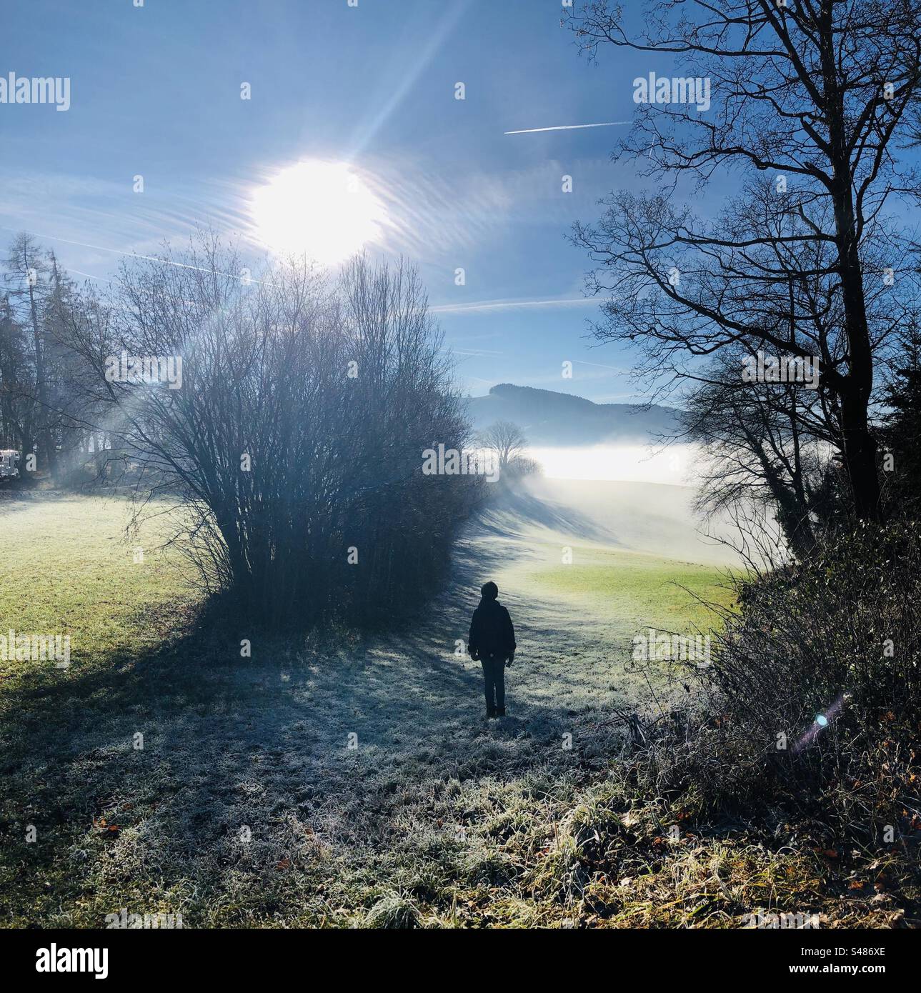 Ein Kind steht mit dem Rücken zu einer seltsamen Landschaft: Der niedrige Nebel verdeckt nicht den blauen Himmel. Die Sonne scheint am blauen Himmel. Das Kind scheint die Elemente zu befehlen. Berge im Hintergrund. Stockfoto