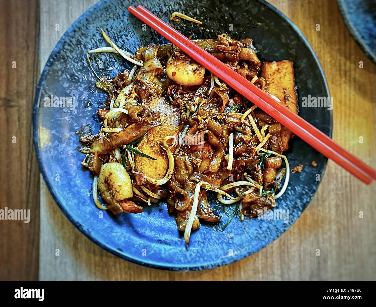 Direkt über dem Blick auf Saibling Koay teow/scharfe, gebratene flache Nudeln mit Garnelen, Mungbohnensprossen und Schnittlauch auf Teller mit roten Stäbchen auf Holztisch. Malaysische/singapurische Küche. - Smartphone-aufgenommenes Stockfoto