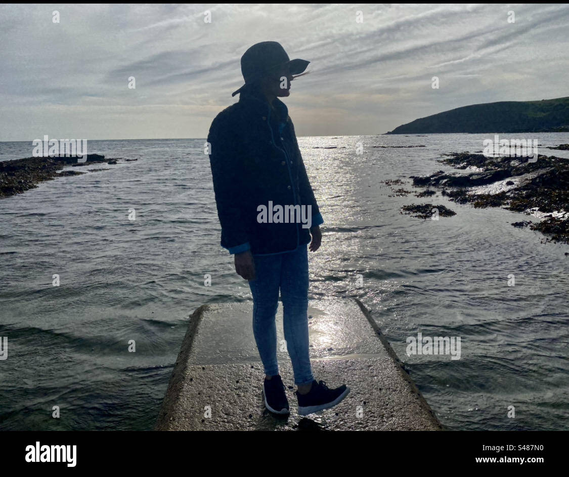 Silhouette mit seitlichem Blick auf das Mädchen mit Hut und Mantel am Meer und Blick über das Wasser - Smartphone-aufgenommenes Stockfoto