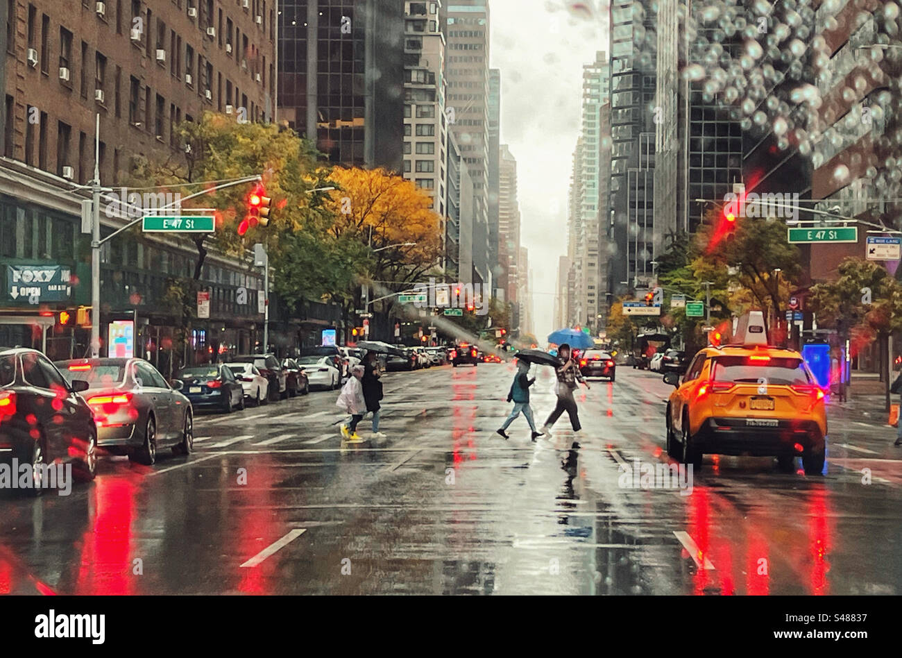 Regentag in New York auf der 47. Straße. Leute auf der Überfahrt mit Sonnenschirmen mit Autos, inklusive Taxi, die warten. Reflexionen auf nasser Straße von Brems- und Bremsleuchten - Smartphone-aufgenommenes Stockfoto