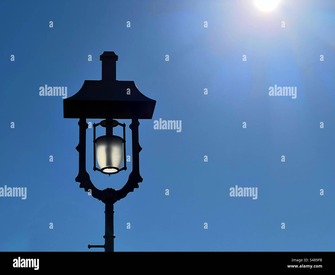 Lampe mit Hintergrundbeleuchtung an der Brooklyn Bridge vor einem leuchtend blauen Himmel mit der Sonne am Rand des Rahmens - Smartphone-aufgenommenes Stockfoto