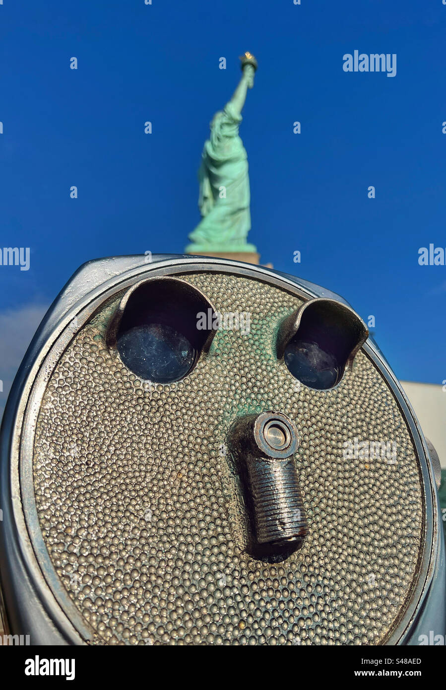 Nahaufnahme des Teleskops auf Liberty Island mit Freiheitsstatue im Hintergrund vor einem leuchtend blauen Himmel - Smartphone-aufgenommenes Stockfoto