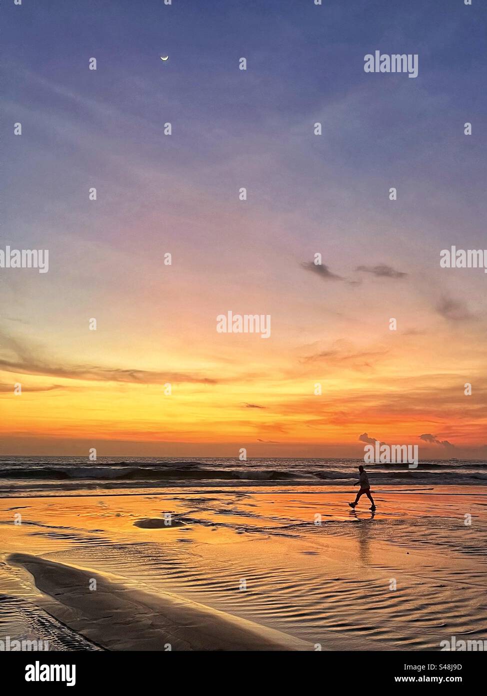 Mann, der bei Sonnenuntergang am Strand in Seminyak Bali spaziert, mit einer Silhouette gegen das Meer und das orange leuchtende Licht am Himmel Stockfoto