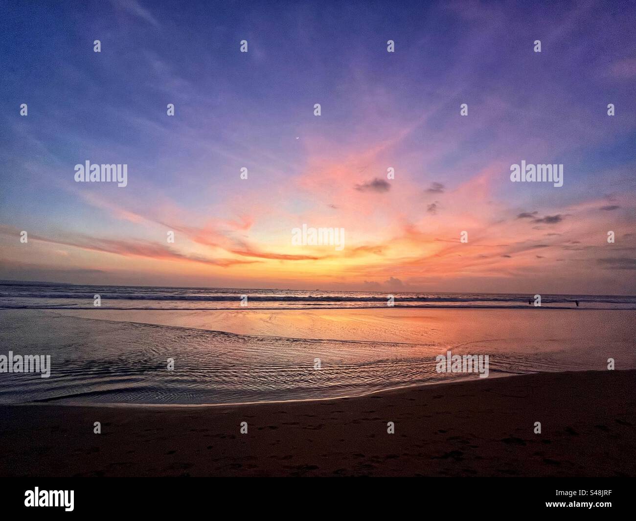 Sonnenuntergang am Seminyak Beach Bali mit dem Sonnenuntergang über dem Meer und dem orange-violetten Leuchten, das in den Wolken verbleibt, und dem orangen Licht, das im flachen Wasser reflektiert - Smartphone-aufgenommenes Stockfoto
