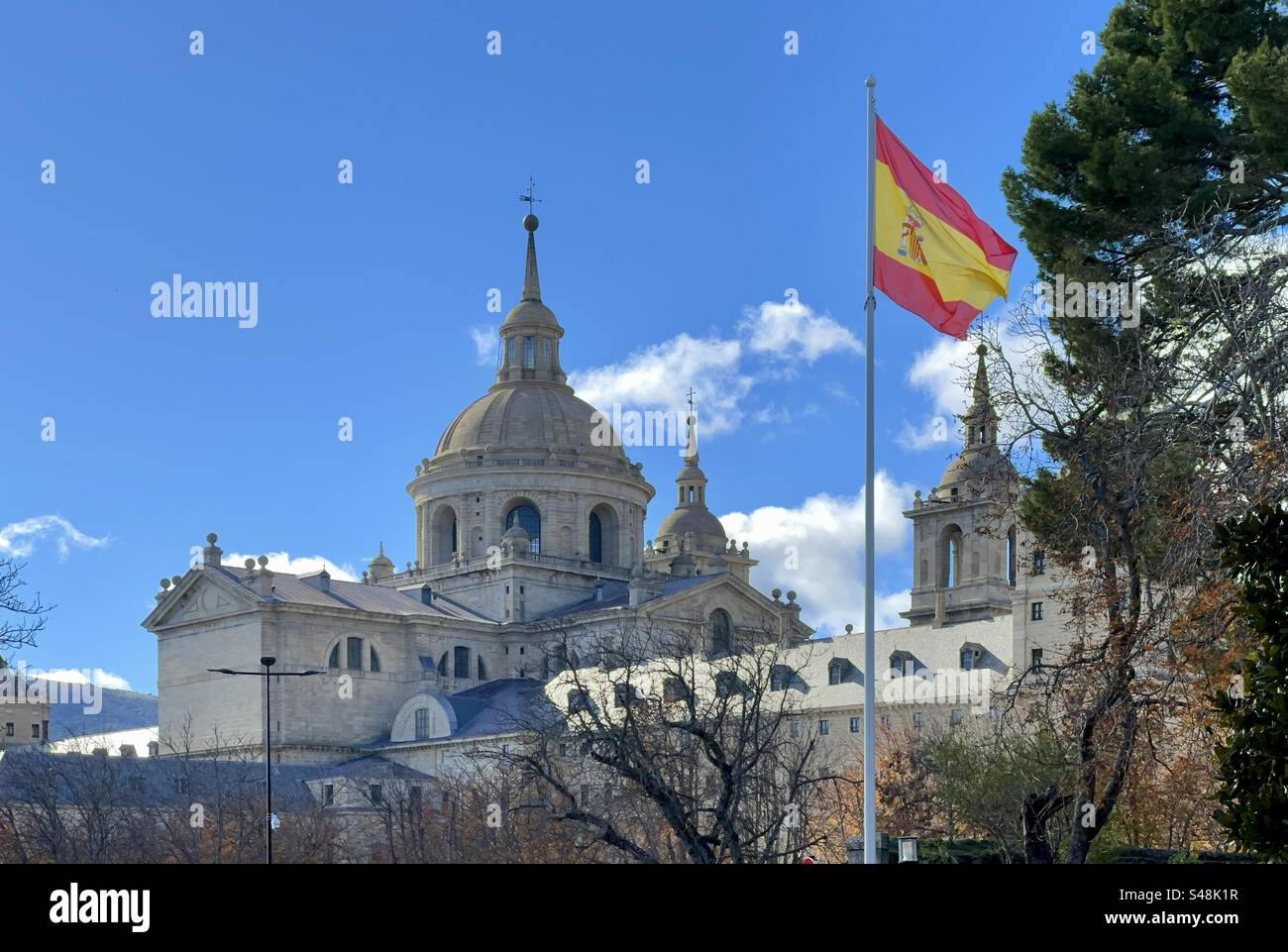 Eine spanische Flagge, die vor dem Königlichen Kloster El Escorial in Madrid, Spanien, fliegt. Ungewöhnliche Ansicht mit Kopierraum am blauen Himmel. Stockfoto
