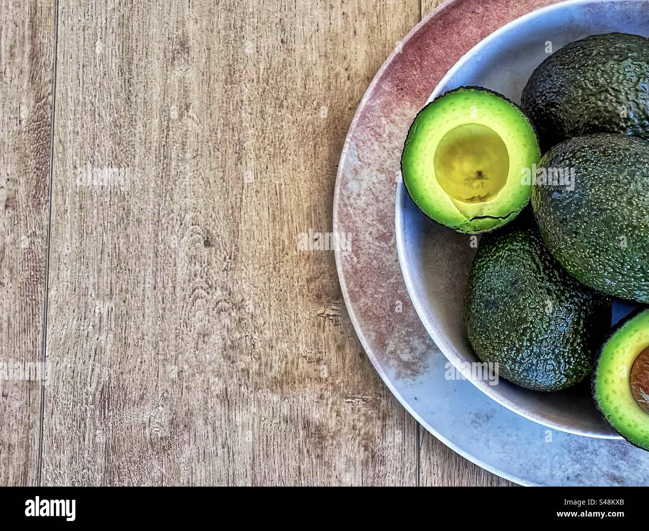 Avocados ganz und in Schüssel auf Teller auf Holztisch mit Kopierraum schneiden. - Smartphone-aufgenommenes Stockfoto