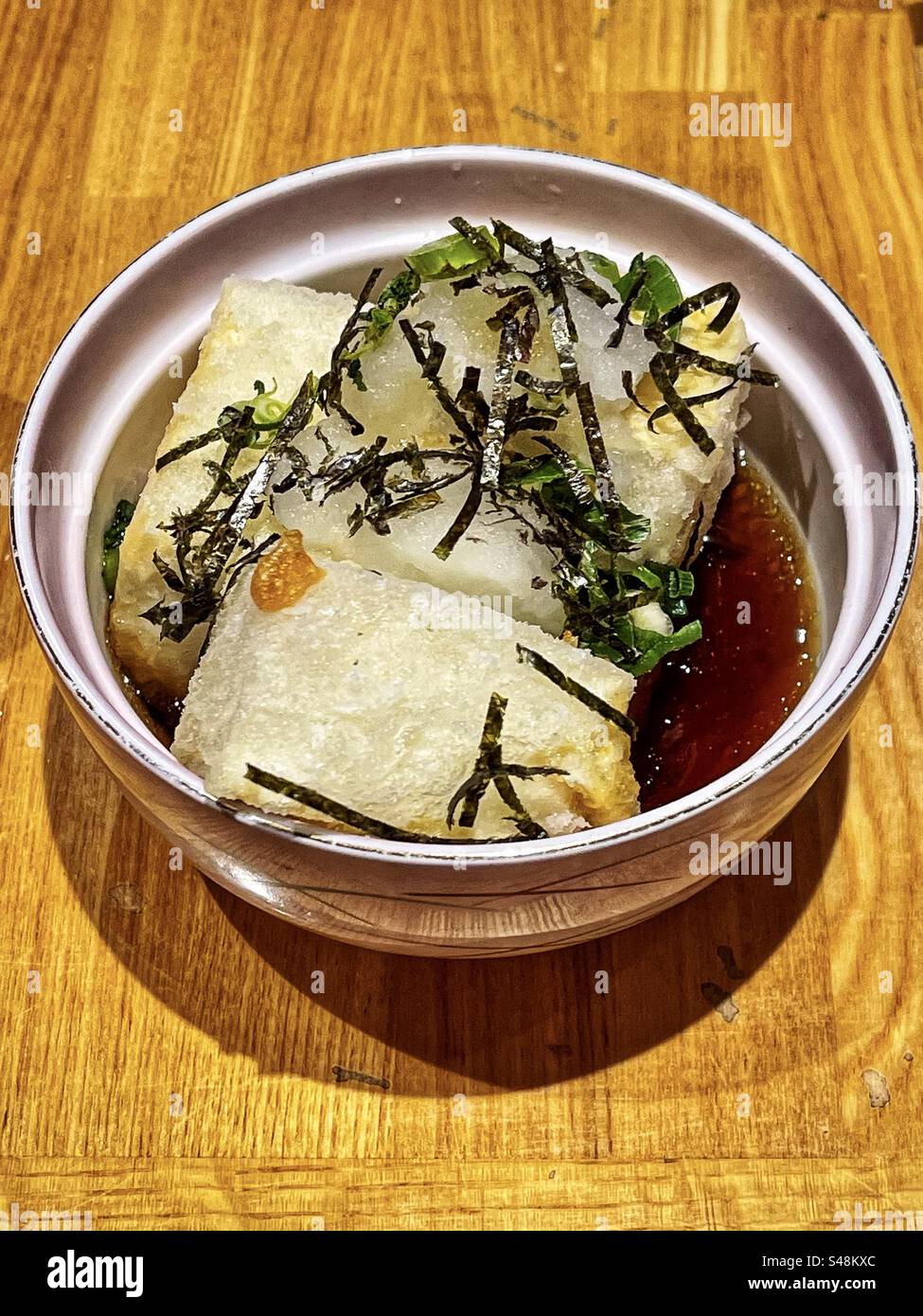 Hochwinkelansicht von Aagedashi Tofu/frittiertem Tofu in heißer Brühe mit Frühlingszwiebeln und geriebenem Daikon in Schüssel auf Holztisch. Japanisches Essen. Vegetarische Speisen. Gesunde Ernährung. Stockfoto