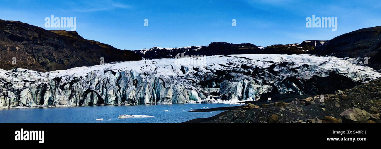 Eisbrocken, die in einer Lagune aus braunem Wasser an einem Gletscher in Island schwimmen. Berge im Hintergrund. Der Himmel ist blau mit Wolken. Das Eis ist blau. Kleine Wellen im Wasser. Schwarze Asche aus dem Vulkan. Stockfoto