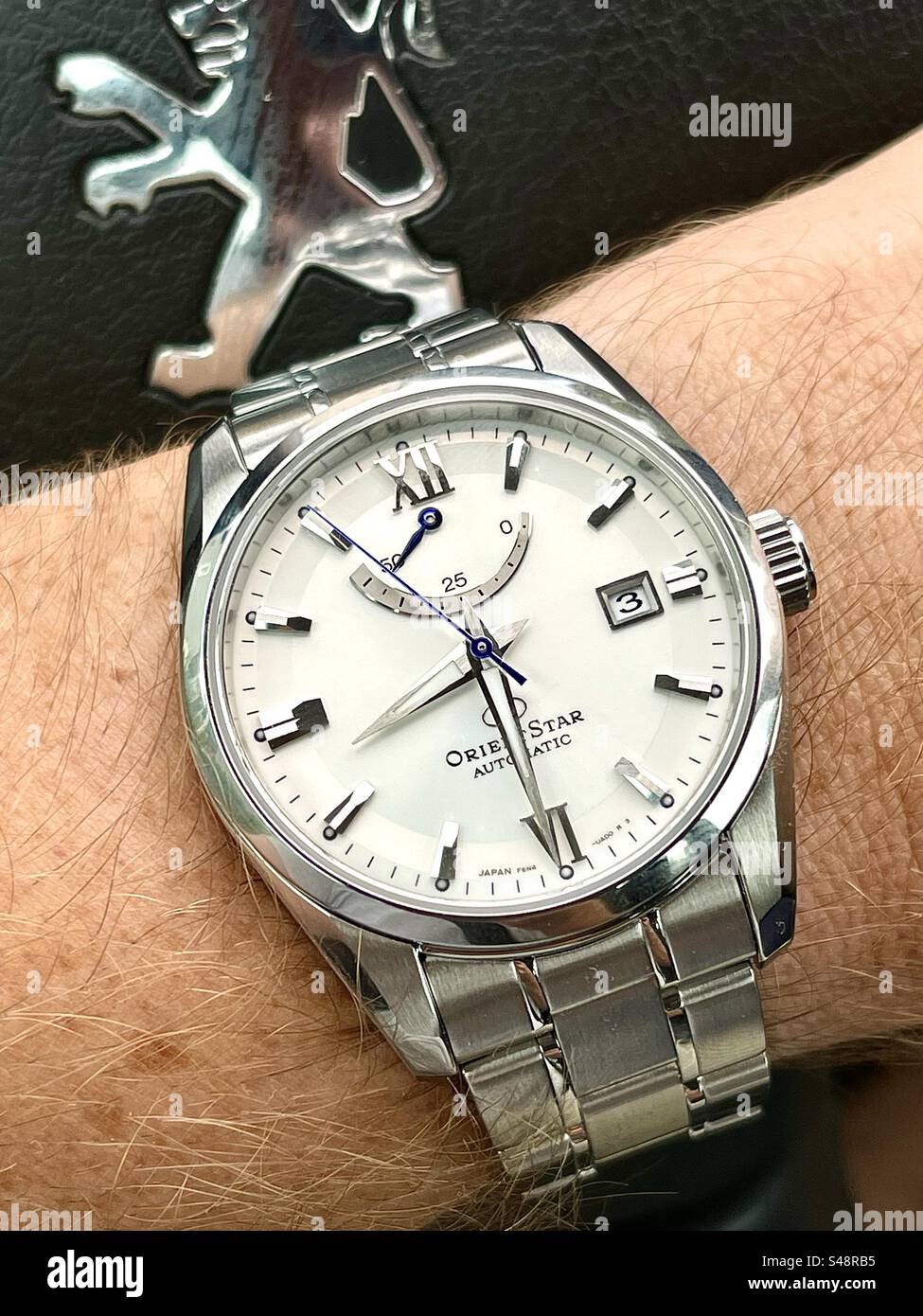 Nahaufnahme einer Orient Contemporary RE-AU0006S00B OrientStar Armbanduhr mit weißem Zifferblatt mit Sonnenschliff und schwarzem Lederhintergrund mit Peugeot-Logo am Lenkrad. Stockfoto