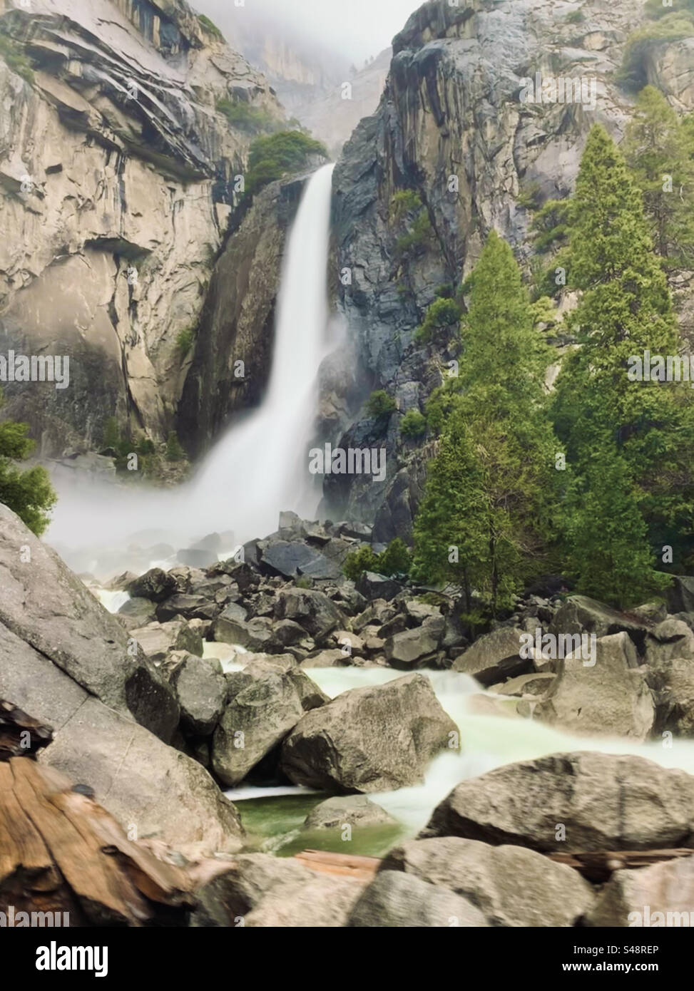 Ein Wasserfall neben einer felsigen, nassen Klippe mit hellgrünen Kiefern auf der rechten Seite. Nebel und Nebel am Himmel. Große Felsen und rauschendes Wasser. Lange Belichtung. Yosemite National Park, Kalifornien. Naturfoto. Stockfoto