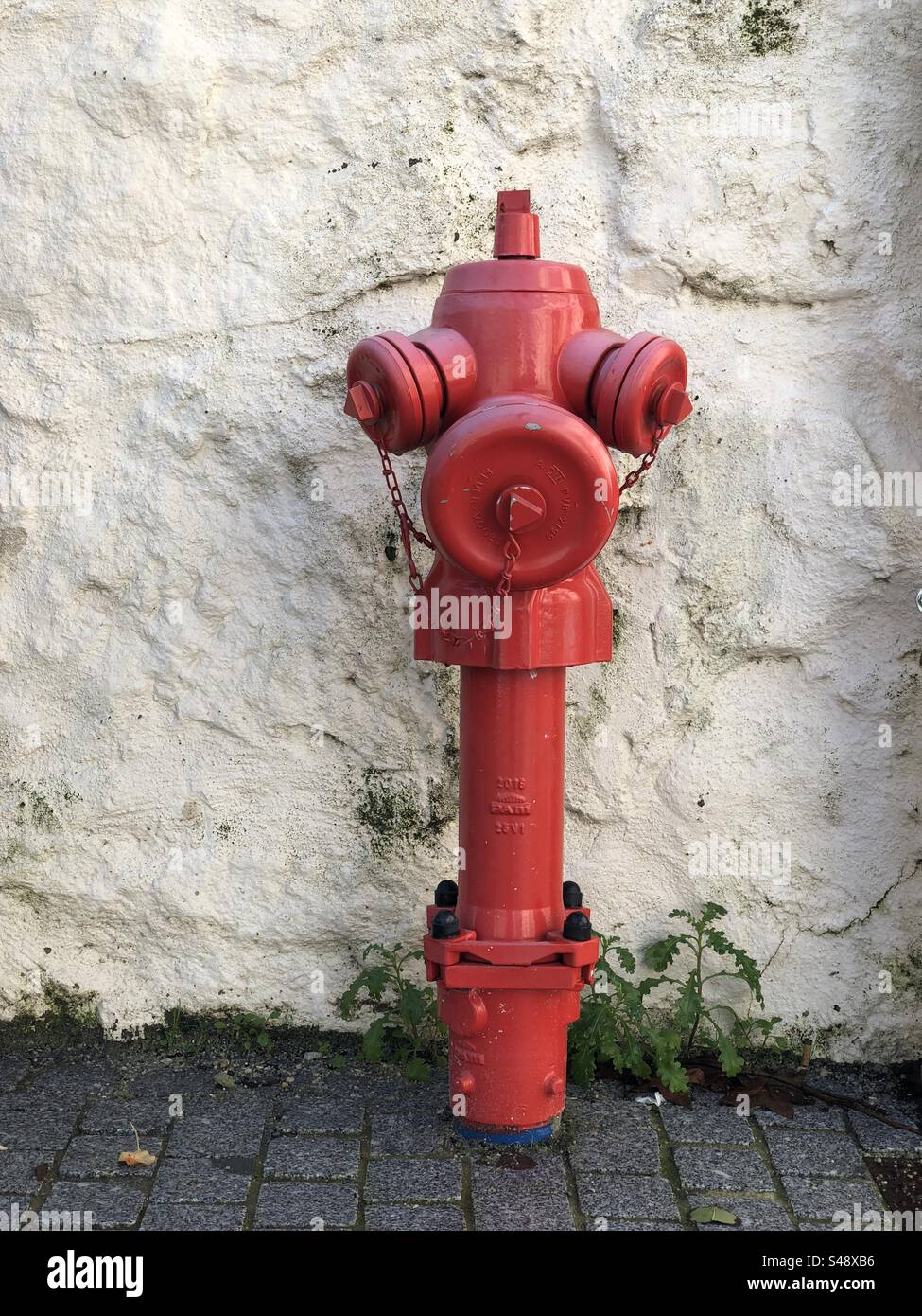 Viana do Castelo, Portugal - 01. Dezember 2023: Ein leuchtender roter Hydrant mit Ketten steht an einer strukturierten weißen Wand auf einer kopfsteingepflasterten Straße Stockfoto