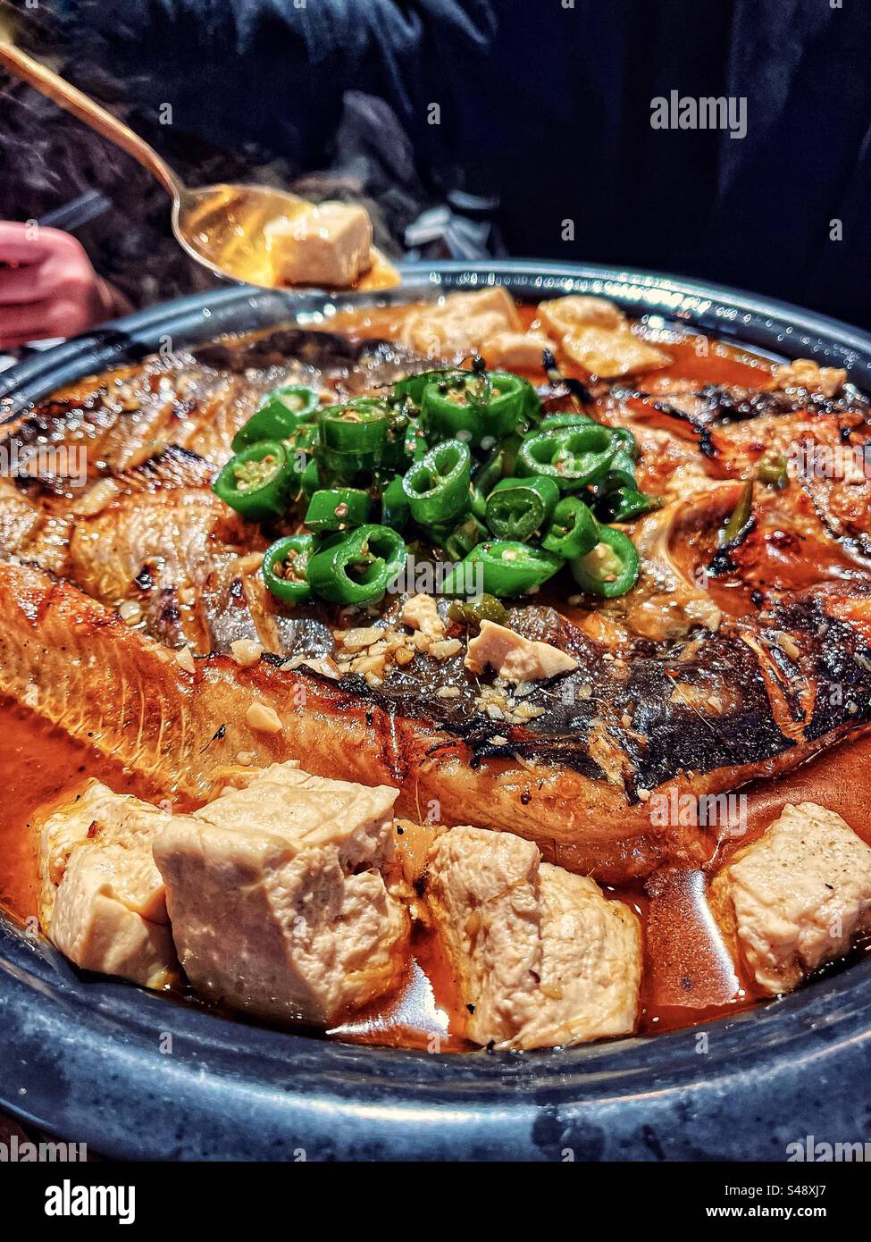 Nahaufnahme von gegrilltem Fisch und Tofuwürfeln in heißer, scharfer Suppe mit grünem Chili im Metalltopf. Chinesische Küche. - Smartphone-aufgenommenes Stockfoto