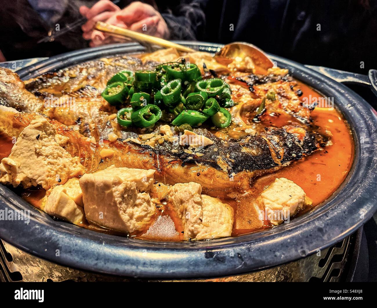 Nahaufnahme von gegrilltem Fisch mit Tofuwürfeln in heißer, scharfer Suppe mit grünem Chili im Metalltopf. Chinesische Küche. - Smartphone-aufgenommenes Stockfoto