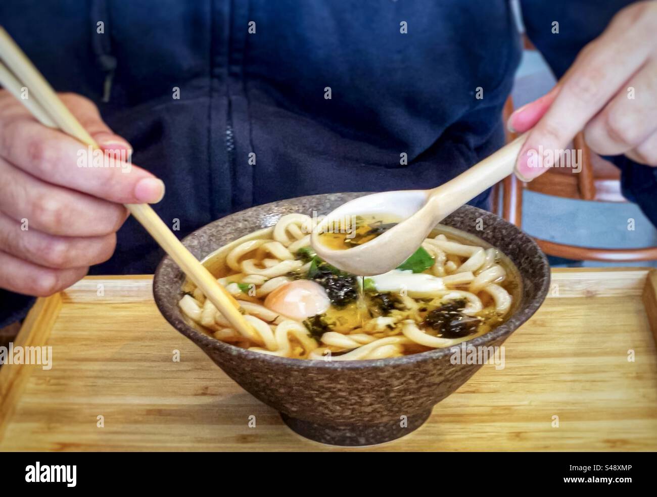 Mittelteil des Mannes mit Essstäbchen und einem Löffel, um Udon-Nudeln in Miso-Brühe mit einem pochierten Ei in einer Schüssel auf Holztablett zu essen. - Smartphone-aufgenommenes Stockfoto