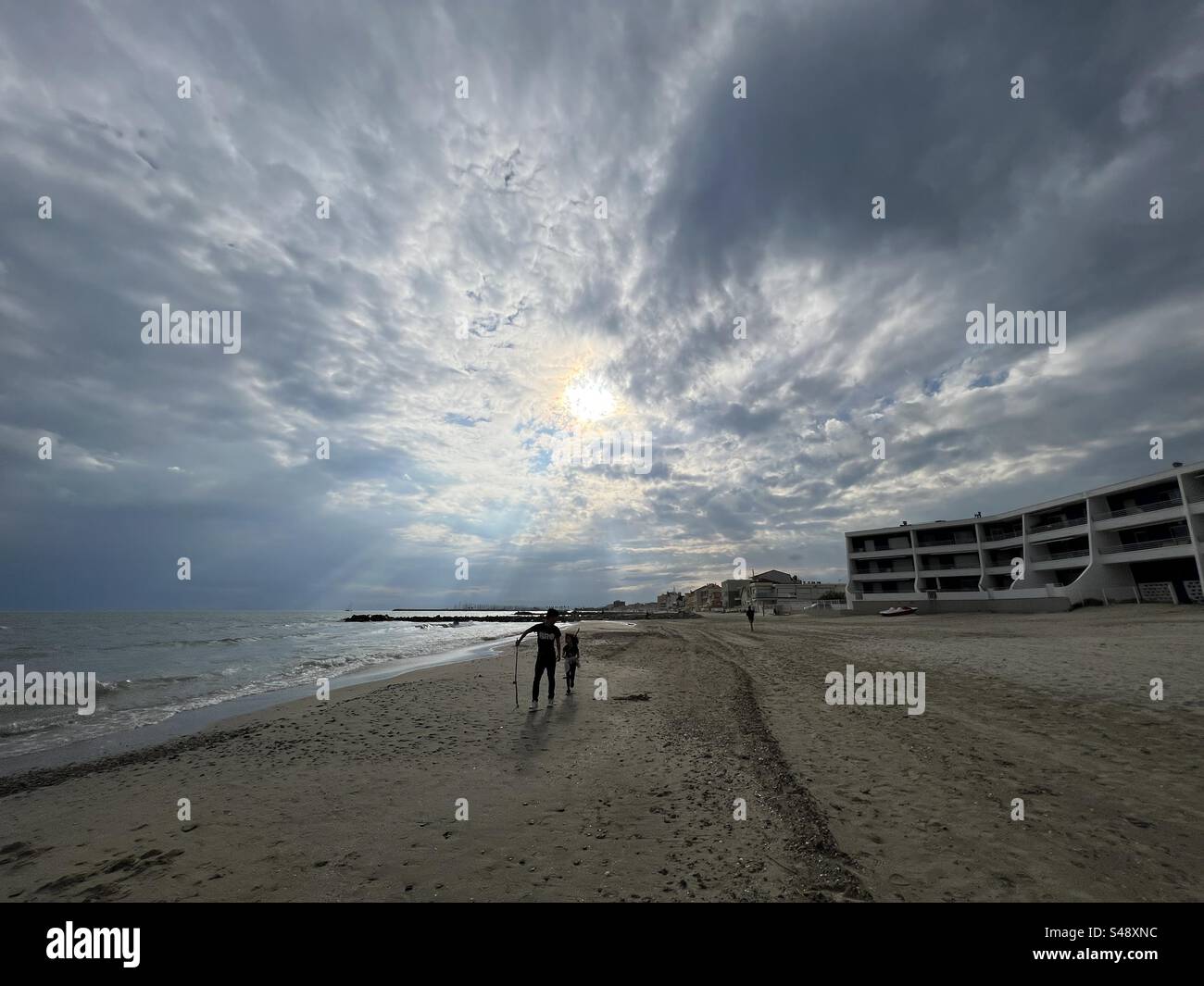 Weite Landschaft eines leeren Strandes mit Sonnenuntergang hinter Wolken in Palavas mit zwei Kindern, großem Bruder und kleiner Schwester, Occitanie, in der Nähe von Montpellier, Frankreich Stockfoto