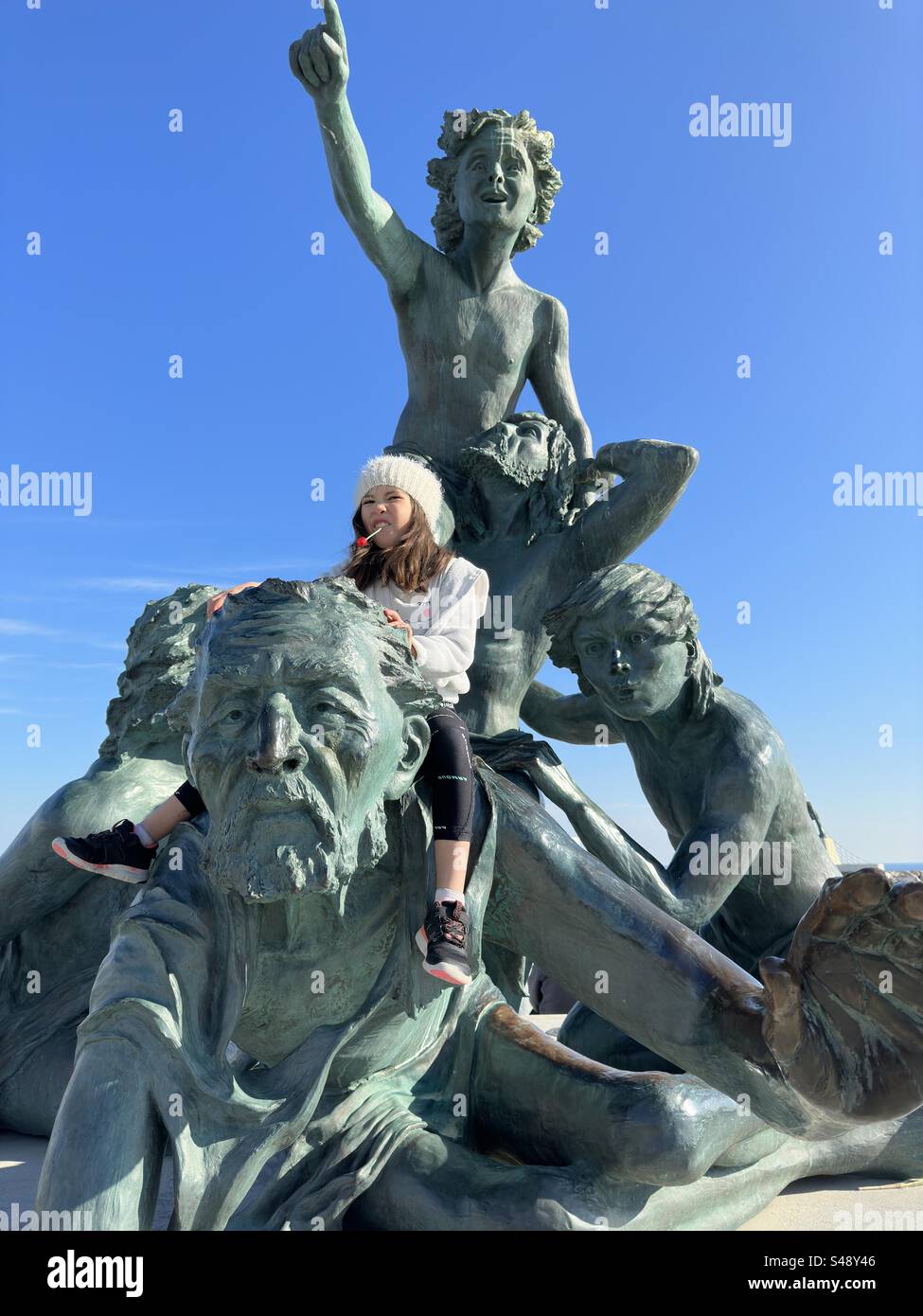 Ein kleines Mädchen mit Winterkleidung und einer weißen Mütze, das an einem Lutscher lutscht, sitzt auf einer Statue in Palavas, Occitanie, Frankreich. Stockfoto