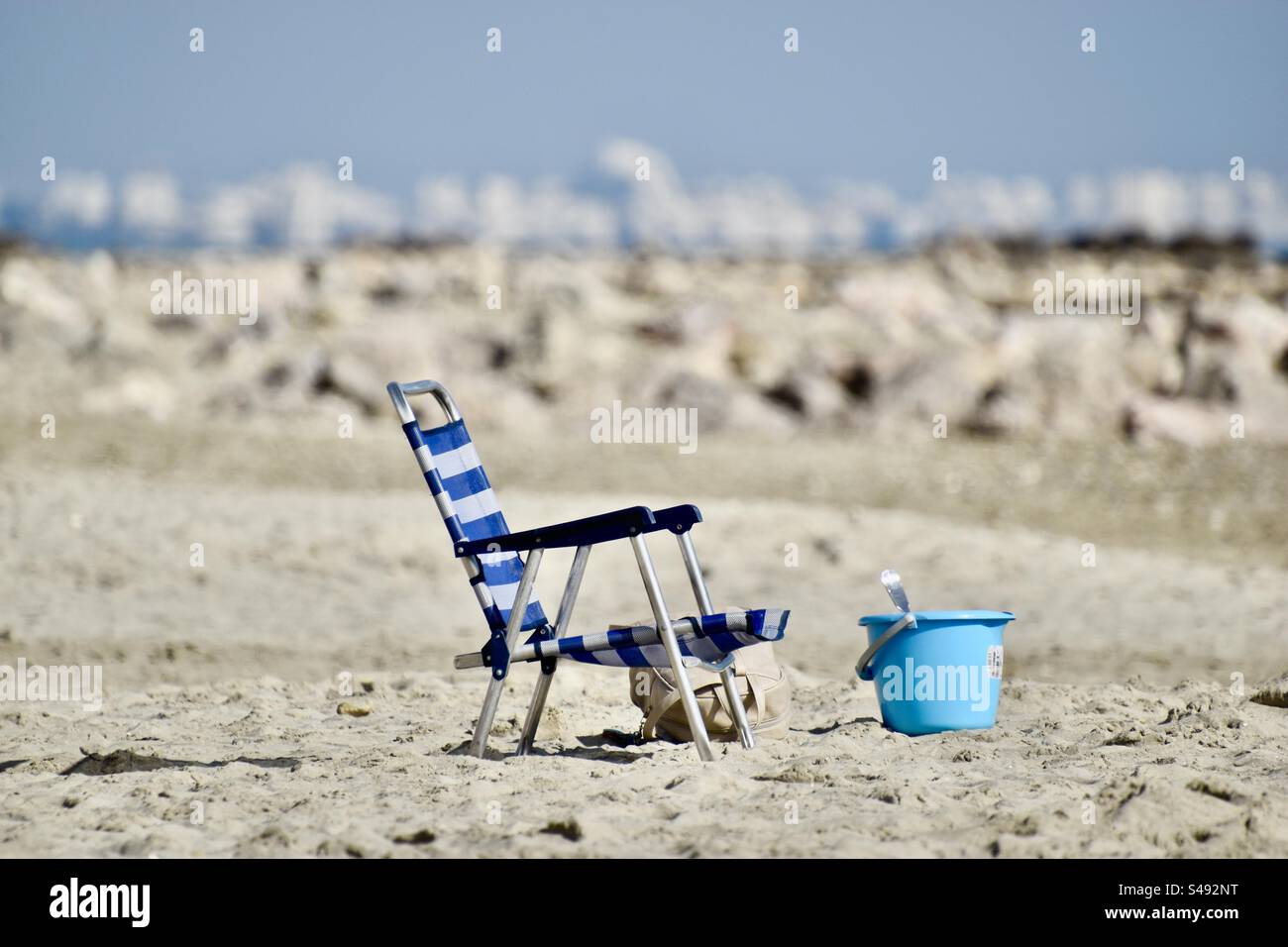 Tele-Aufnahme eines leeren, blau-weißen, klappbaren Liegestuhls mit Strandspielzeug in Palavas, Occitanie, Frankreich während des Sommers. - Smartphone-aufgenommenes Stockfoto