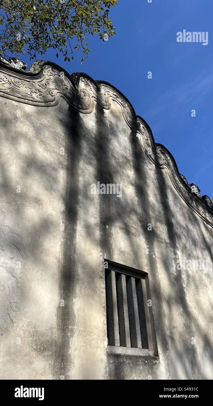 Eine traditionelle chinesische Gebäudewand mit einer einzigartigen wellenförmigen Dachlinie und einem Fenster, die weiche Schatten von den umliegenden Bäumen wirft. Stockfoto