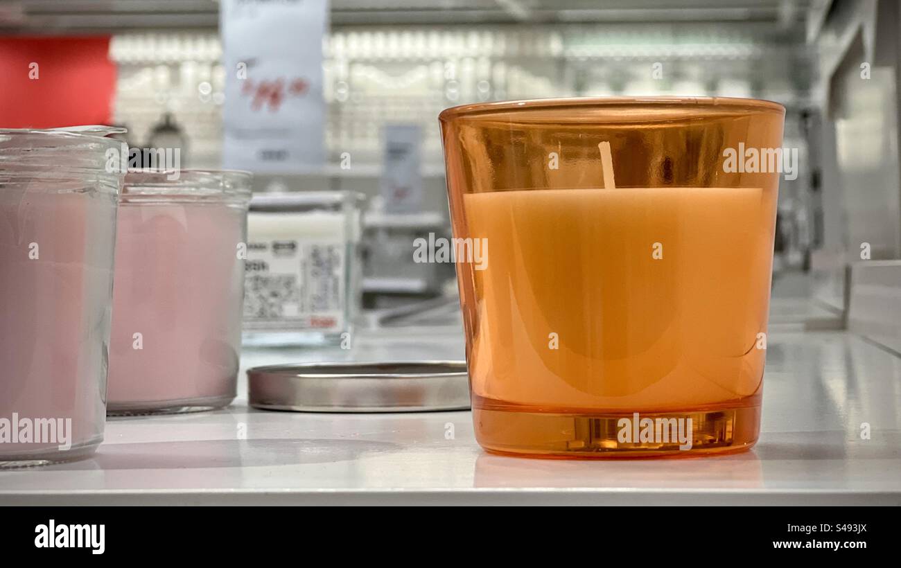 Nahaufnahme einer weißen Kerze in einem orangefarbenen Glashalter. Flacher Fokus mit Verkaufsregalen im Hintergrund Stockfoto