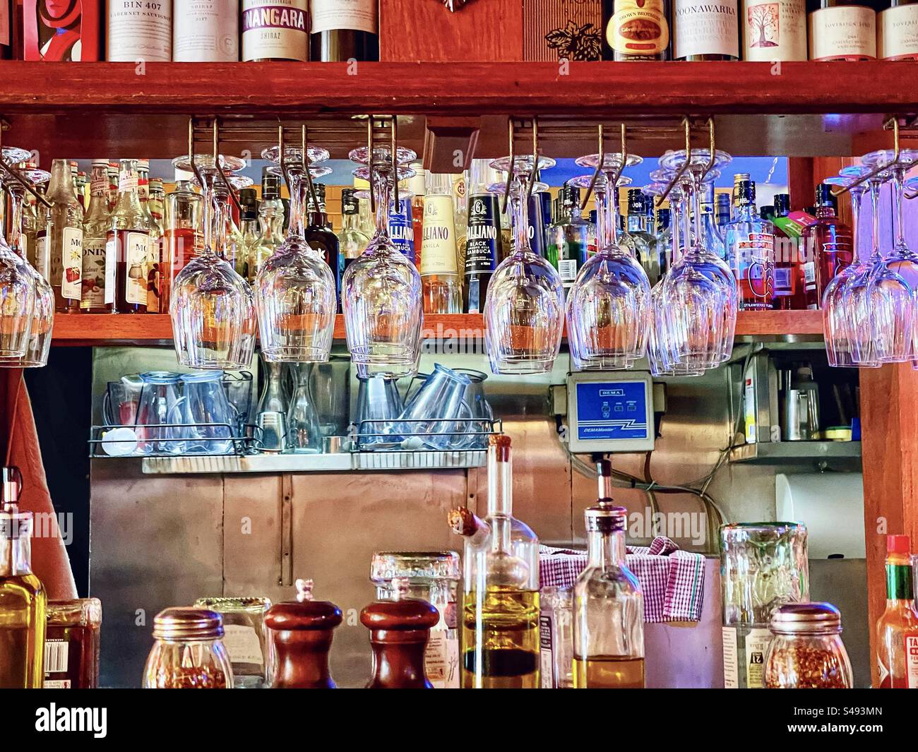 Leere Weingläser hängen an den Regalen eines italienischen Restaurants mit Gewürzen und Olivenölflaschen vor Flaschen mit Hartalkohol. Stockfoto