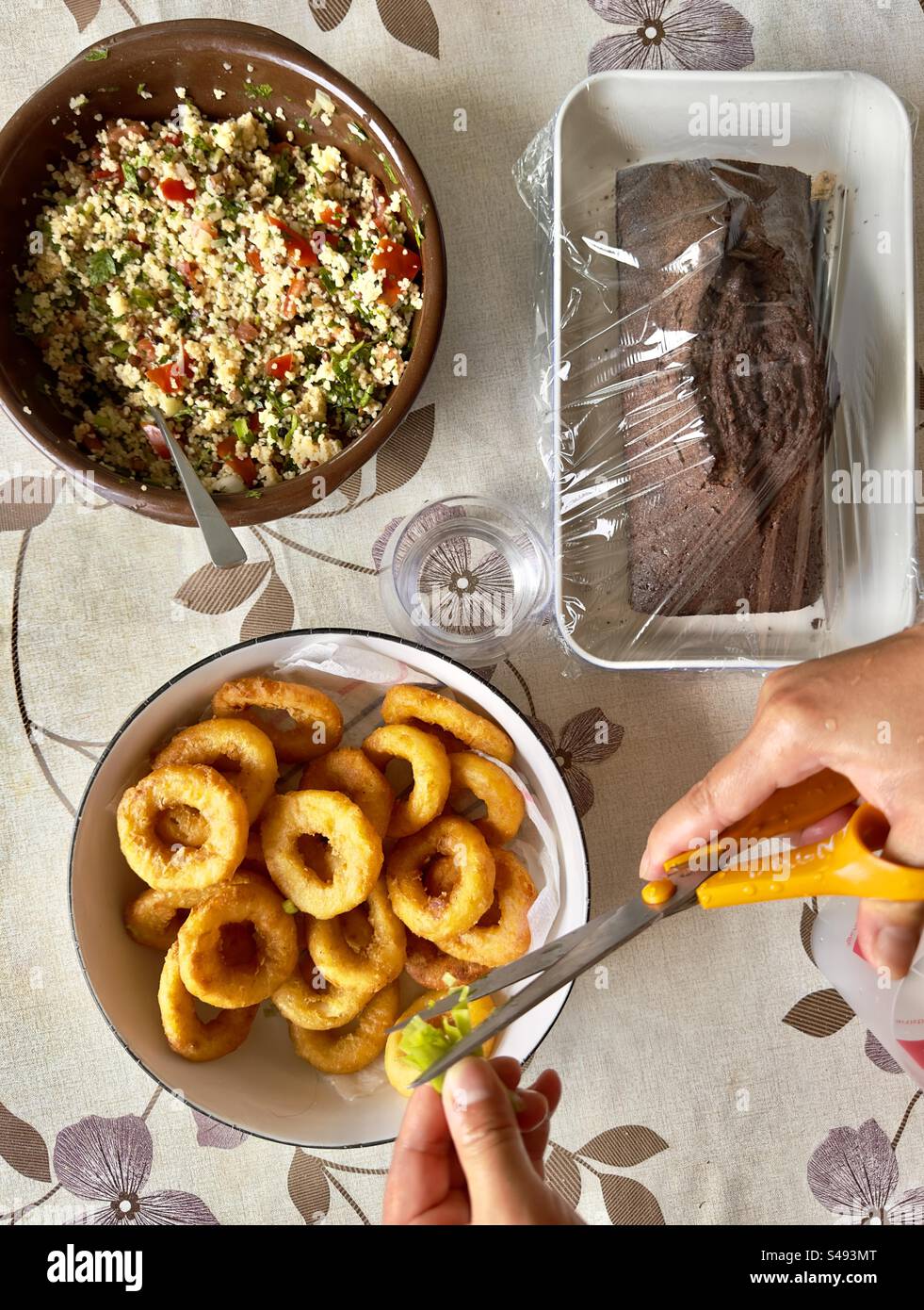Top-down-Aufnahme eines Küchentisches mit drei Gerichten: Ein Taboulé, Zwiebelringe und ein Schokoladenmarmorkuchen. Hände einer jungen Asiatin schneiden grüne Zwiebeln über die Ringe. - Smartphone-aufgenommenes Stockfoto