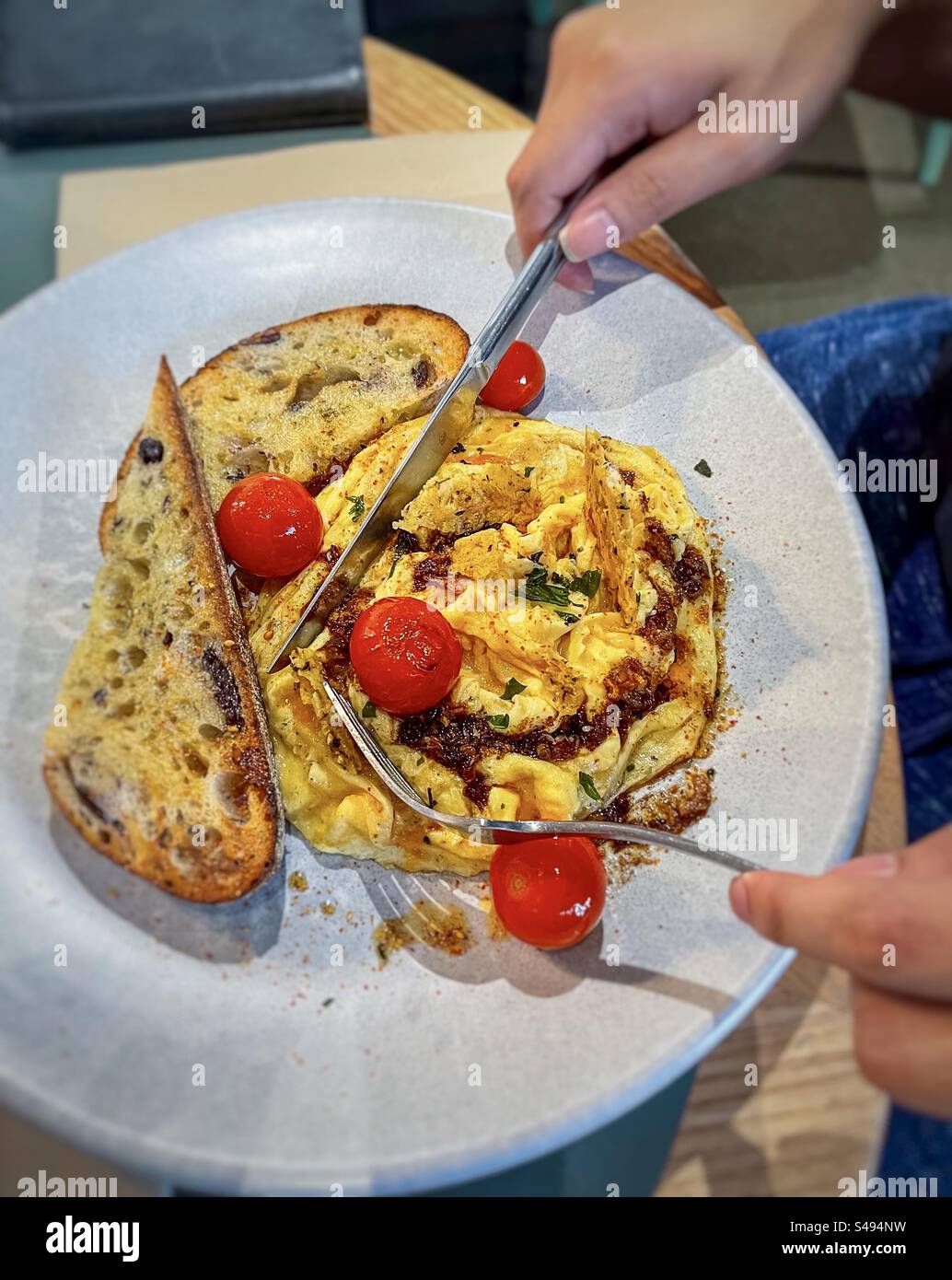 Hände halten Messer und Gabel über Chili Rührei mit Kirschtomaten und Sauerteig Toast auf Teller auf dem Tisch. Gesundes Frühstück. Stockfoto