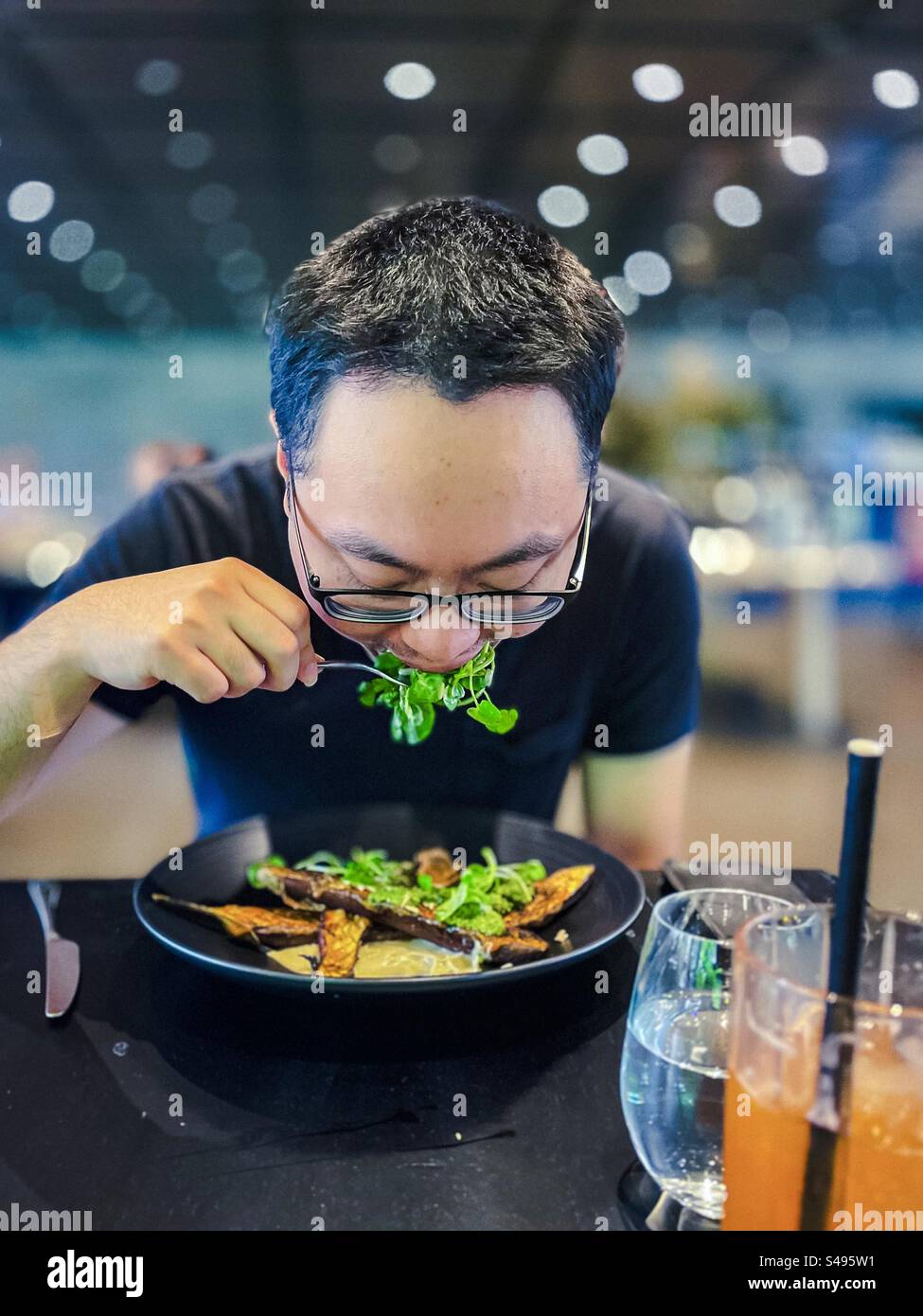 Ein Mann in Brille, der eine Mahlzeit aus frischem Salat und Miso marinierten, gerösteten Auberginen am Tisch im Restaurant genießt. Gesunde Ernährung. Vegetarische Mahlzeit. Fokus auf den Vordergrund. Stockfoto