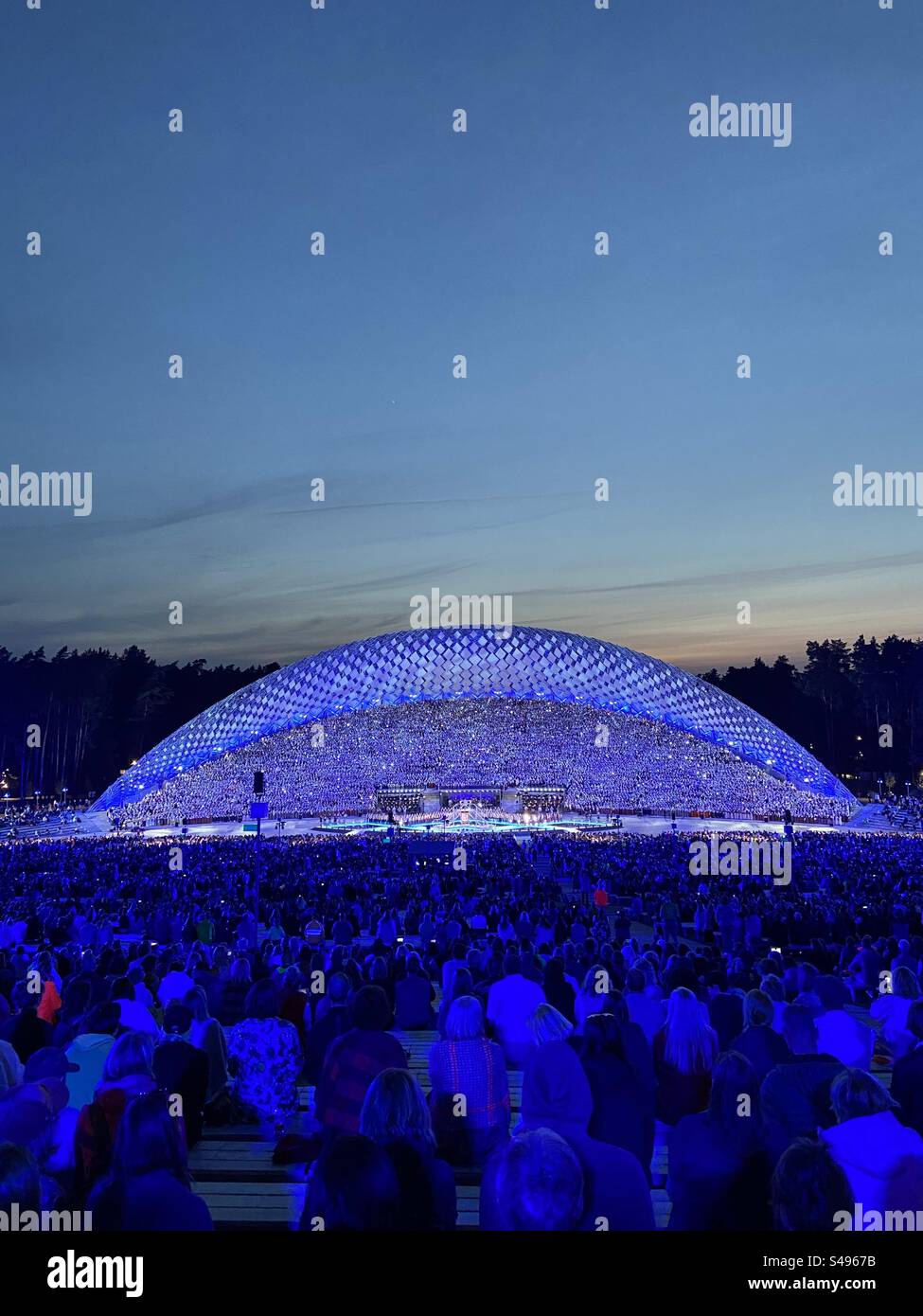 Leute, die das Konzert des lettischen Gesangs und Tanzfestivals am Veranstaltungsort des Großen Gesangs in Riga, Lettland, beobachten. Viele blaue Lichter und Lichteffekte auf der Bühne. Foto aufgenommen, wenn es dunkel wird Stockfoto