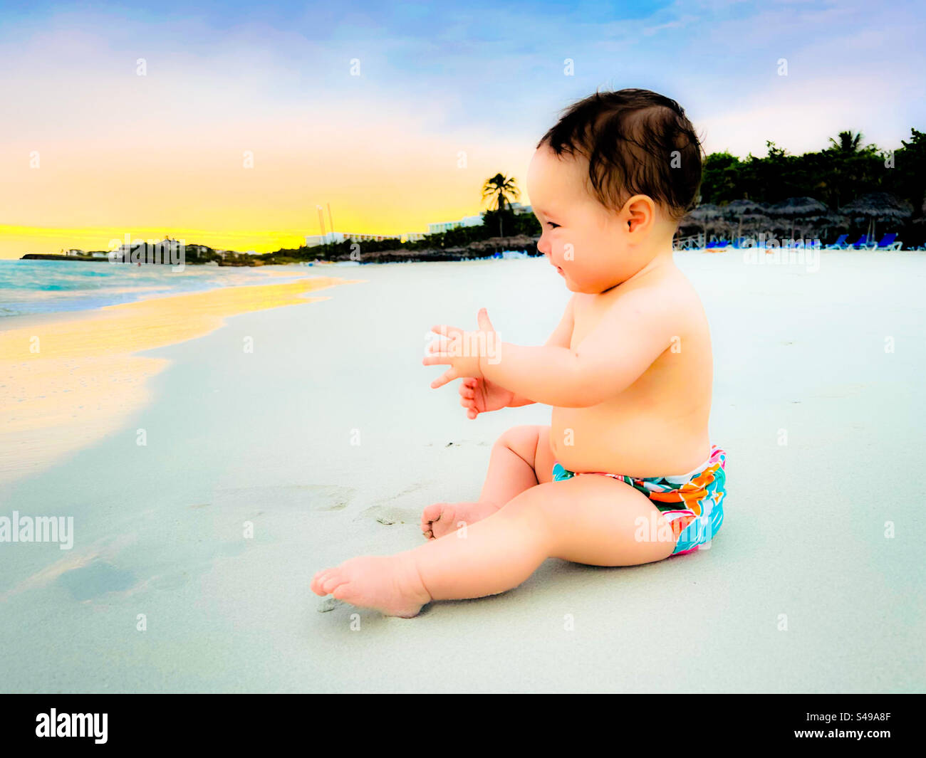 Strandausflug mit Baby. Glückliches Baby, das auf dem Sand sitzt und auf das Meer blickt. Varadero, Kuba. Träumerischer Filter - Smartphone-aufgenommenes Stockfoto