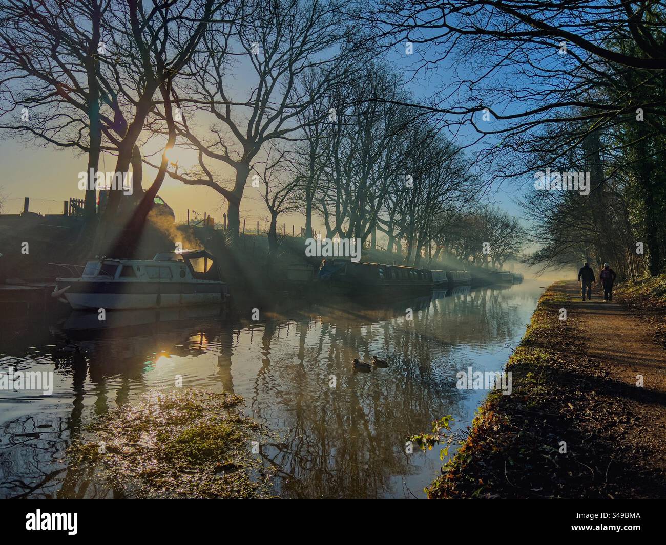 Reflexionen im Morgennebel des Winters auf dem Leeds- und Liverpool-Kanal in Adlington, Lancashire. Zwei Personen laufen auf einem Schleppweg mit Reflexen von Bäumen und schmalen Booten im Kanal. Stockfoto