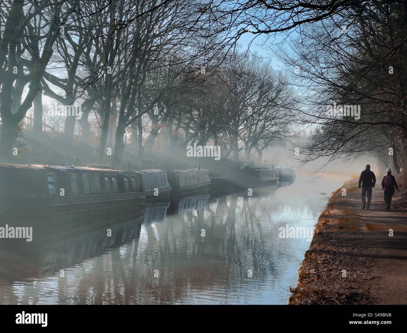 Zwei Personen laufen auf dem Schleppweg von Leeds und Liverpool Kanal im Morgennebel. Schmale Boote und Bäume spiegeln sich im Kanal. Adlington bei Chorley in Lancashire. Stockfoto