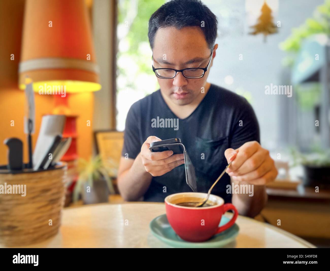 Asiatischer Mann mit Brille, der ein Handy benutzt, während er eine Tasse heißen schwarzen Kaffee am Tisch im Café rührt. Selektiver Fokus. Stockfoto