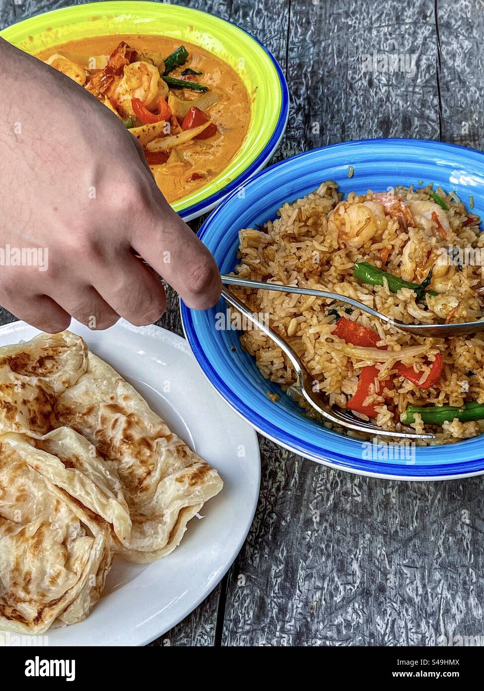 Löffel und Gabel mit der Hand über gebratenem thailändischem Basilikumreis in einer Schüssel mit rotem thailändischem Curry in einer Schüssel und Roti, gebratenem Fladenbrot auf dem Teller auf dem Tisch. Thailändisches Essen. - Smartphone-aufgenommenes Stockfoto