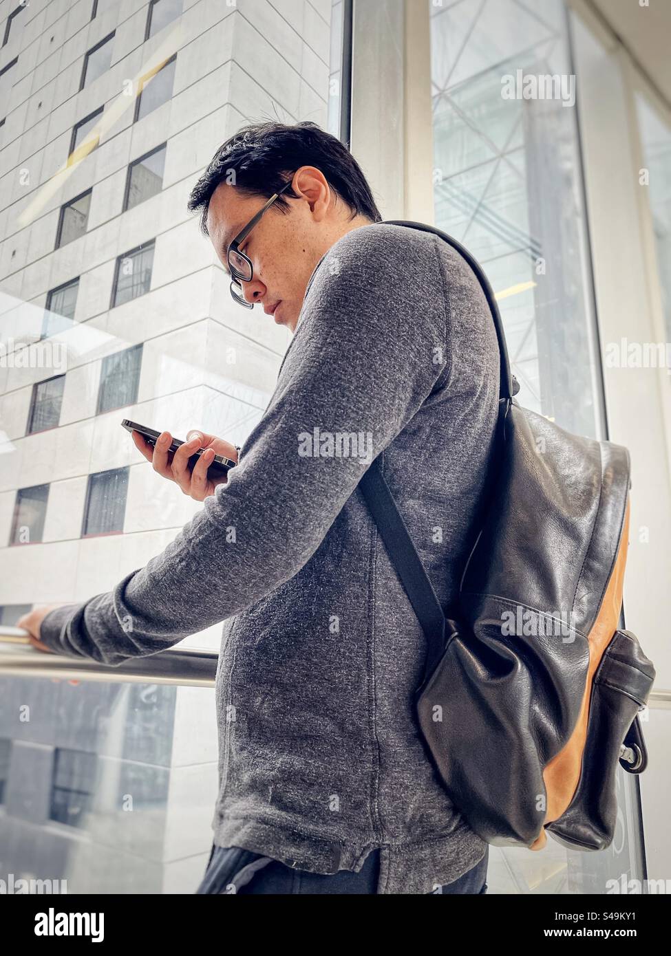 Tiefwinkelansicht eines asiatischen Mannes mit Rucksack mit Handy gegen Fensteransicht der Gebäude in der Stadt. - Smartphone-aufgenommenes Stockfoto