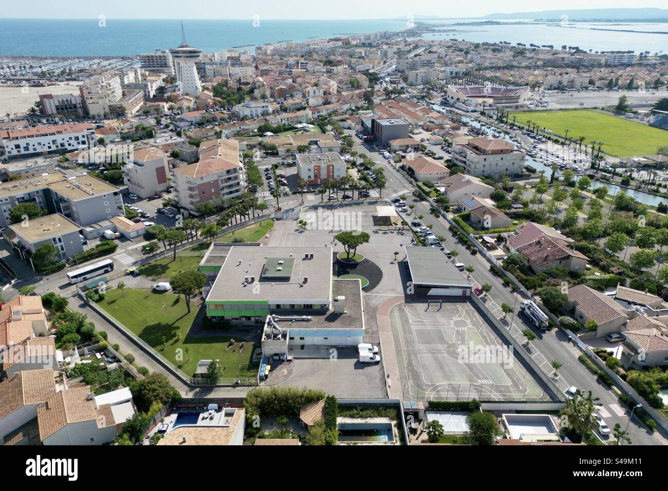 Drohnenaufnahme der öffentlichen Schule in Palavas-les-Flots mit der Innenstadt, Tour de La Méditerranée und dem Mittelmeer als Kulisse. Stockfoto